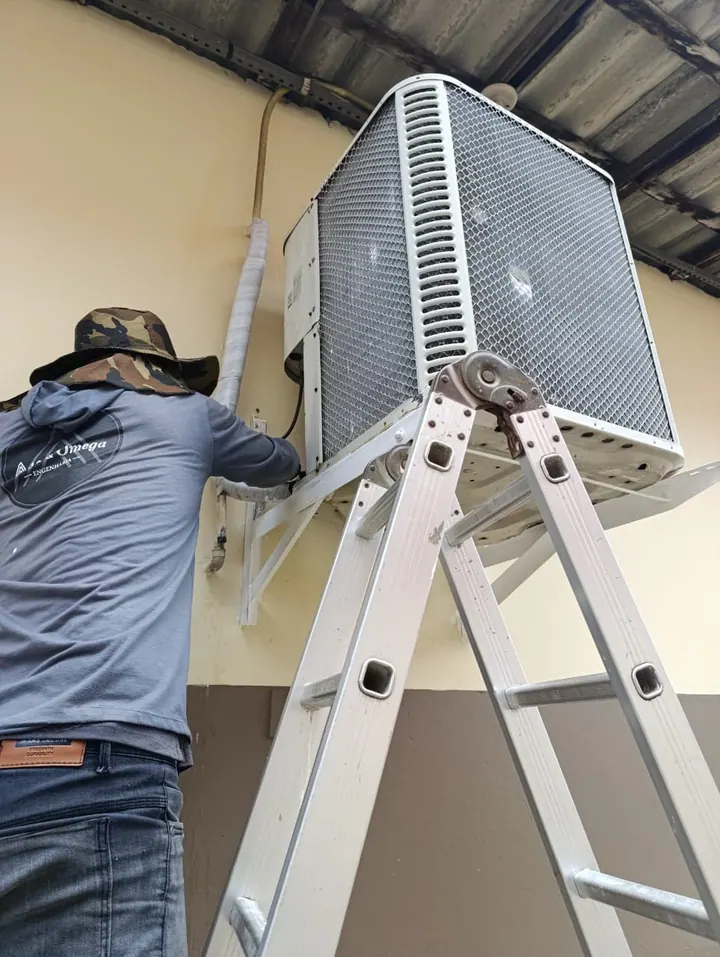 Técnico da Alfa & Ômega em escada fixando uma condensadora de ar-condicionado em suporte na parede